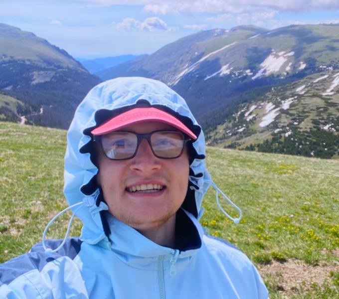 A man in a light blue jacket with the hood up, a red hat brim, on a mountain
